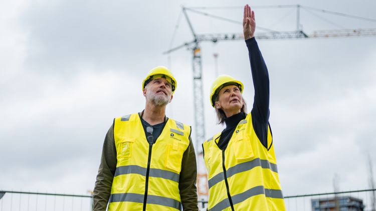 Bilde av en byggeplass, en mann og en kvinne peker på noe i luften. De har på vernehjelm og refleksvester fra Arbeidstilsynet