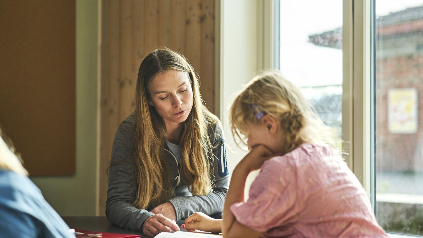 En lærer sitter ved et bord sammen med en elev i et klasserom og hjelper henne med en oppgave. En annen elev er delvis synlig i forgrunnen.