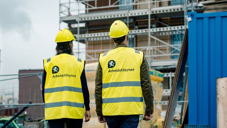 Bildet viser to inspektører med Arbeidstilsynet-vest som står foran en byggeplass.